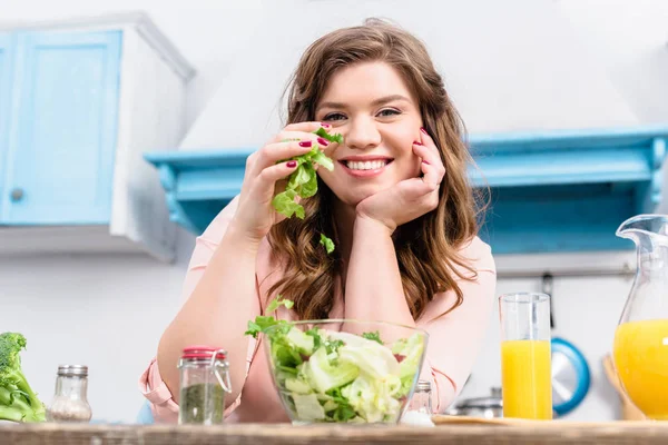 Übergewichtige lächelnde Frau am Tisch mit frischem Salat in der heimischen Küche — Stockfoto