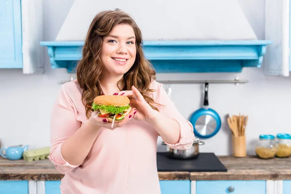 Ritratto di giovane donna sovrappeso con hamburger in mano in cucina a casa — Foto stock
