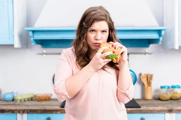 Portrait de jeune femme en surpoids avec hamburger dans les mains dans la cuisine à la maison — Photo de stock