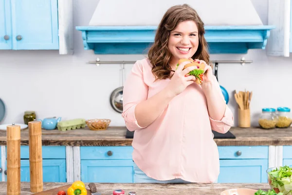 Porträt einer übergewichtigen lächelnden Frau mit Burger in der Hand in der heimischen Küche — Stockfoto