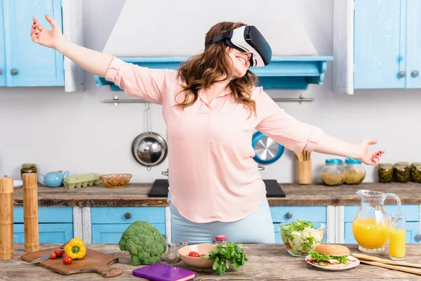 Sonriente mujer con sobrepeso en realidad virtual auriculares de pie en la mesa con verduras frescas en la cocina en casa - foto de stock