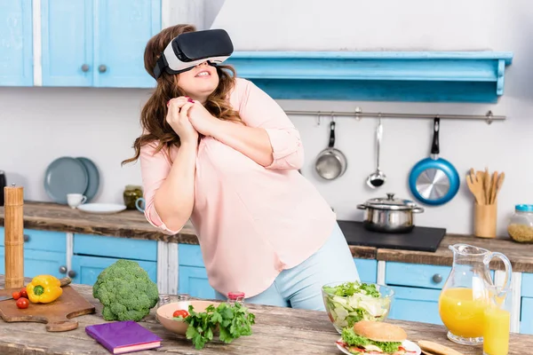 Femme en surpoids effrayée dans le casque de réalité virtuelle debout à table avec des légumes frais dans la cuisine à la maison — Photo de stock