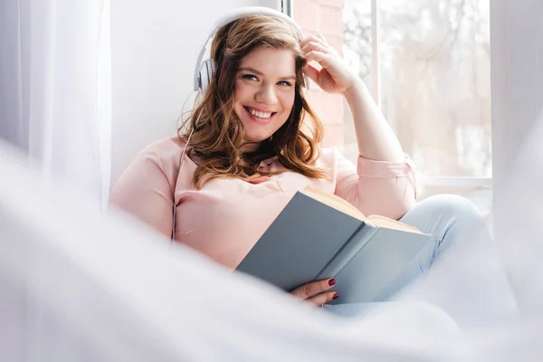 Улыбающаяся женщина в наушниках с книгой, сидящей дома на подоконнике — стоковое фото