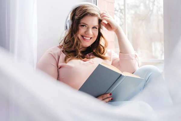 Femme souriante dans un casque avec un livre assis sur le rebord de la fenêtre à la maison — Photo de stock