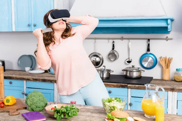 Молода жінка з ножем у гарнітурі віртуальної реальності стоїть за столом зі свіжими овочами на кухні вдома — стокове фото