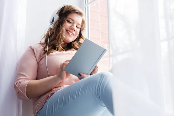 Giovane donna in cuffia leggere libro mentre seduto sul davanzale della finestra a casa — Foto stock