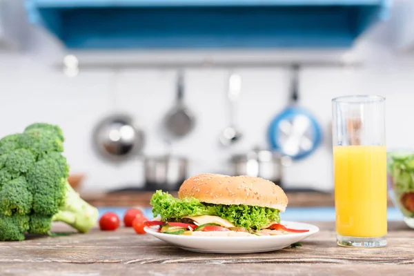 Вид на бургер на тарелке, свежий брокколи и стакан сока на деревянном столе на кухне — стоковое фото