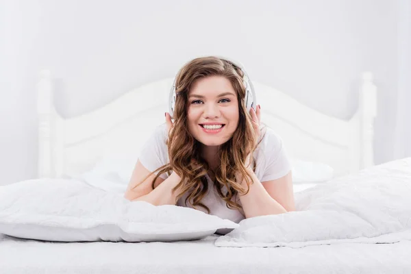 Retrato de jovem mulher sorridente no pijama ouvindo música em fones de ouvido na cama em casa — Fotografia de Stock