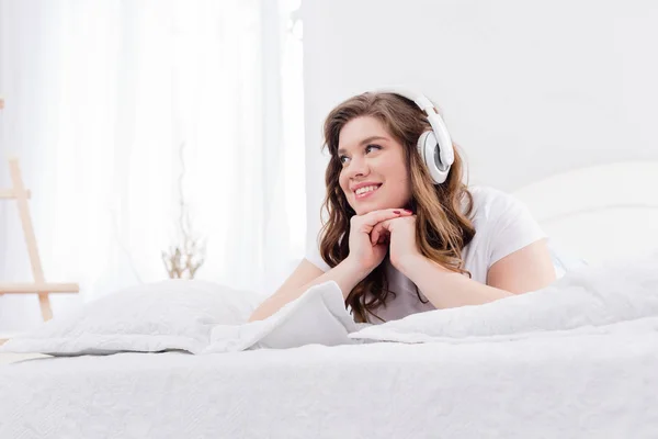 Smiling woman in pajama listening music in headphones on bed at home — Stock Photo