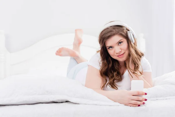 Portrait de jeune femme en pyjama et écouteurs avec smartphone couché sur le lit à la maison — Photo de stock