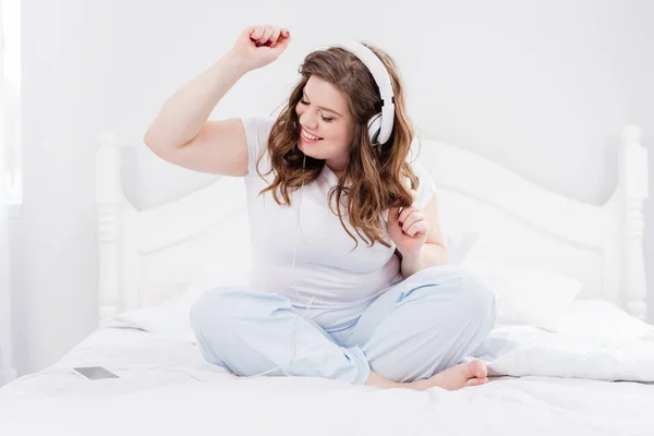 Femme souriante en pyjama écouter de la musique dans les écouteurs sur le lit à la maison — Photo de stock