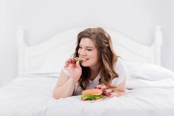 Overweight woman in pajama eating burger on bed at home — Stock Photo