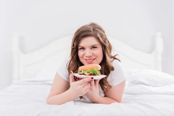 Portrait d'une femme souriante en pyjama avec hamburger sur le lit à la maison — Photo de stock