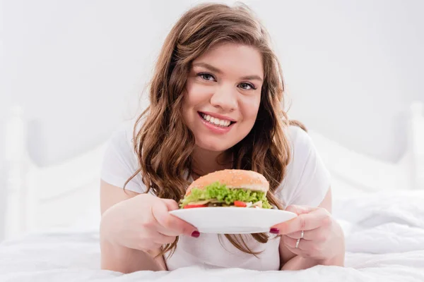 Retrato de mulher sorrindo sobrepeso no pijama com hambúrguer na cama em casa — Fotografia de Stock