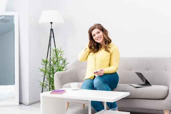 Молодая улыбающаяся женщина со смартфоном сидит на диване с ноутбуком дома — стоковое фото
