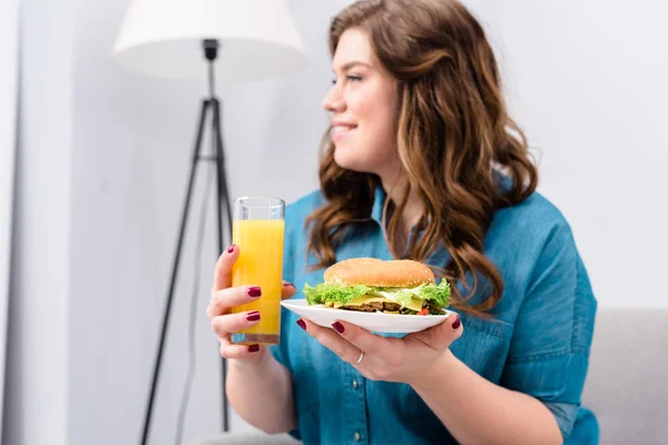 Mulher sorrindo sobrepeso com copo de suco e hambúrguer na placa nas mãos em casa — Fotografia de Stock