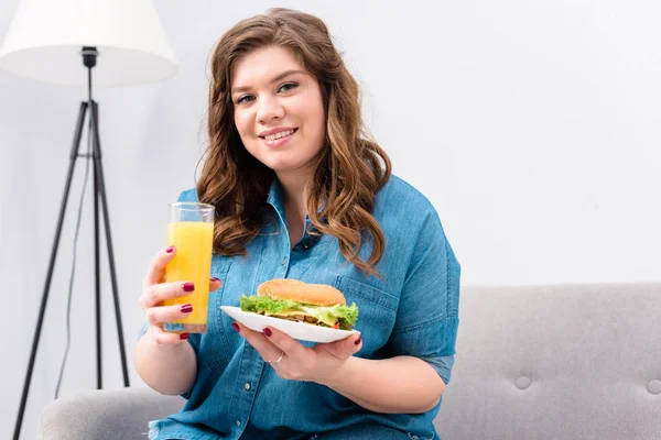 Mulher sorrindo sobrepeso com copo de suco e hambúrguer na placa nas mãos em casa — Fotografia de Stock