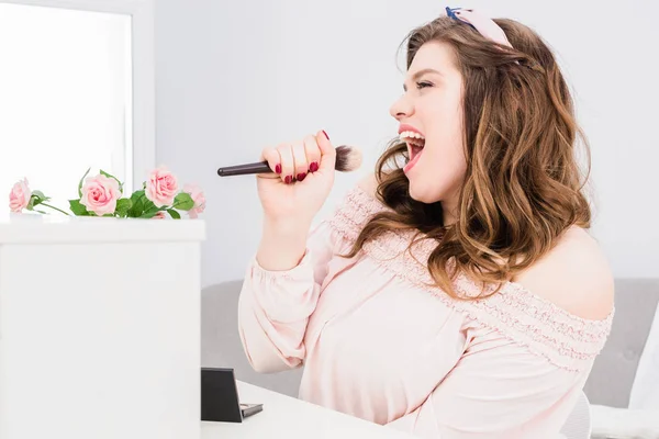 Emotivo giovane donna che canta con pennello trucco in mano come microfono a casa — Foto stock