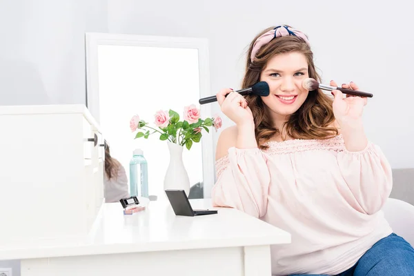 Attrayant jeune femme avec brosses de maquillage dans les mains en regardant la caméra à la maison — Photo de stock
