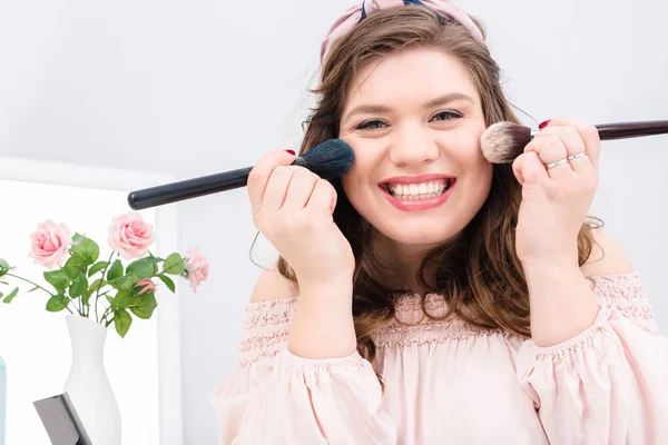 Attraktive junge Frau mit Schminkpinseln in den Händen, die zu Hause in die Kamera schaut — Stockfoto