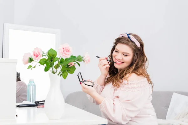 Красивая улыбающаяся молодая женщина наносит блеск для губ, делая макияж дома — стоковое фото
