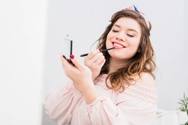 Retrato de mujer joven bastante sonriente aplicando brillo de labios mientras hace maquillaje en casa - foto de stock