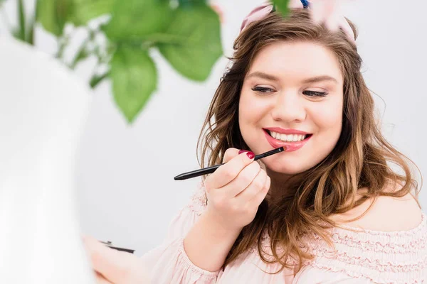 Porträt einer lächelnden jungen Frau, die Lipgloss aufträgt, während sie sich zu Hause schminkt — Stockfoto