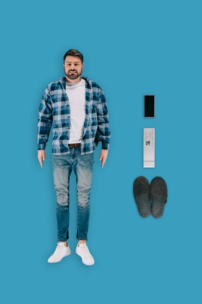 Vista superior del hombre desempleado con smartphone, mando a distancia y zapatillas aisladas sobre fondo azul - foto de stock