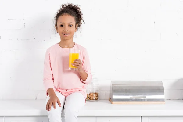 Niño afroamericano con vaso de jugo de naranja sentado en la cocina y mirando a la cámara - foto de stock