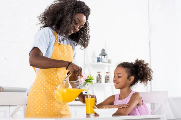 Afro-américaine mère et fille dans des tabliers avec du jus d'orange sur la cuisine — Photo de stock