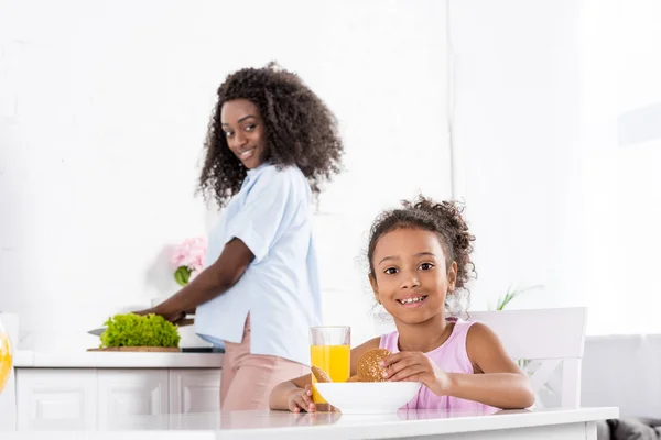 African american daughter with orange juice and cookies on kitchen with mother — Stock Photo