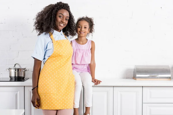 Afroamericana madre en delantal e hija posando en cocina - foto de stock