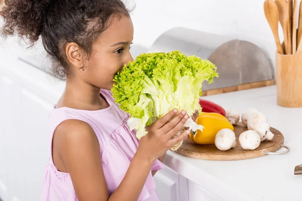 Criança afro-americana farejando alface verde na cozinha — Fotografia de Stock