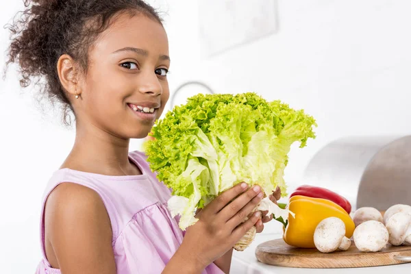 Sorrindo criança afro-americana segurando alface verde fresca na cozinha — Fotografia de Stock