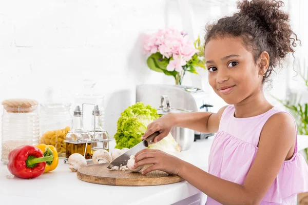 Bambino africano americano che taglia verdure su tavola di legno e guarda la macchina fotografica — Foto stock