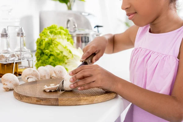 Cropped view of african american kid cutting mushrooms on wooden board in kitchen — Stock Photo