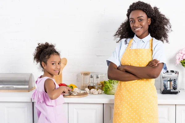 Madre afroamericana con brazos cruzados e hija cortando verduras en la cocina - foto de stock