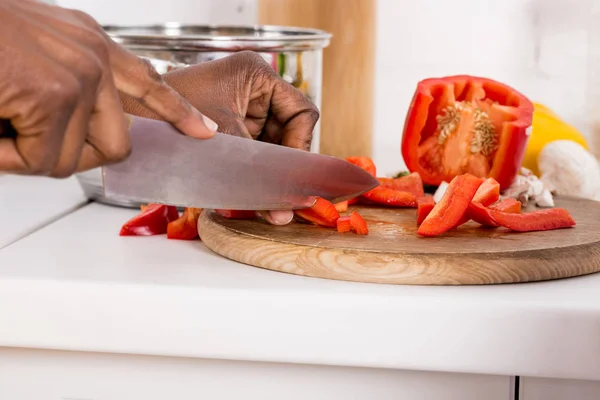 Vista cortada da mulher americana africana cortando pimentão vermelho na placa de madeira na cozinha — Fotografia de Stock