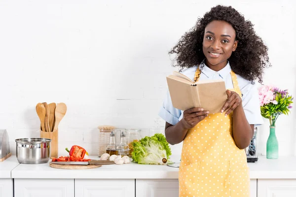 Sorrindo mulher americana africana no avental leitura livro de receitas na cozinha — Fotografia de Stock