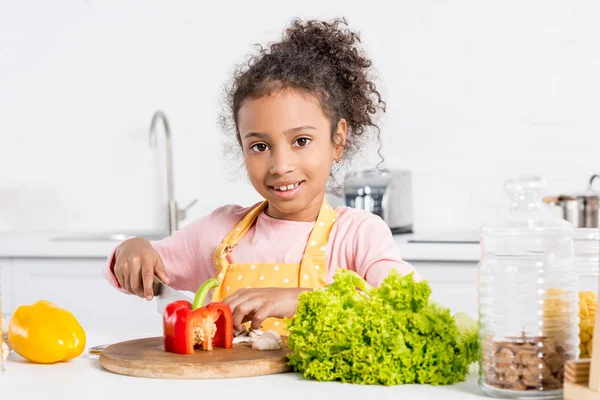 Adorable african american kid in apron cutting bell peppers and lettuce on wooden board in kitchen — Stock Photo