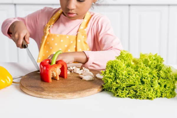 Vue recadrée de gamin afro-américain dans tablier coupe légumes sur planche en bois dans la cuisine — Photo de stock