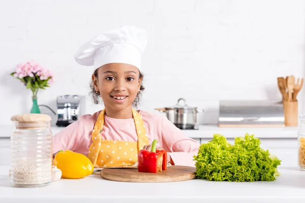 Niño afroamericano sonriente en delantal y sombrero de chef preparando verduras en la cocina - foto de stock