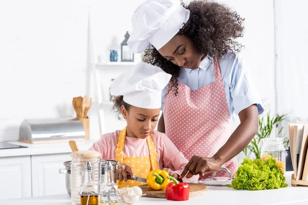 Africano americano madre e hija en chef sombreros cortar verduras en cocina - foto de stock