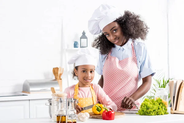 Happy african american mother and daughter in chef hats cutting bell pepper on kitchen — Stock Photo