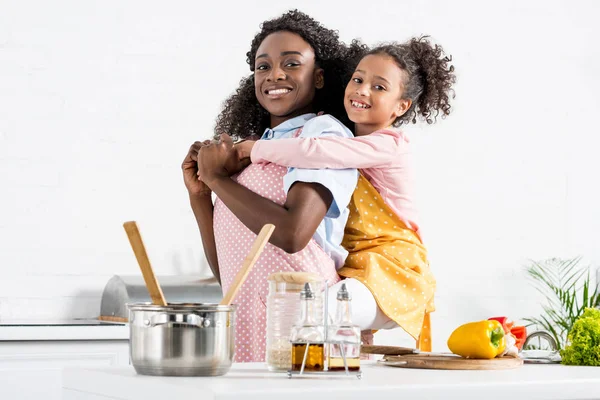 Feliz africano americano madre piggybacking hija en cocina - foto de stock