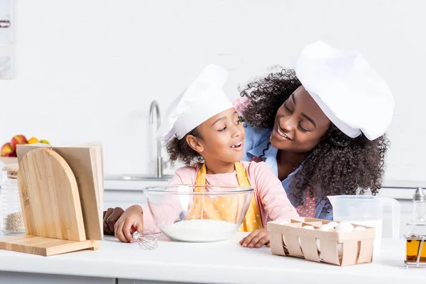 Heureuse mère afro-américaine et sa fille dans des chapeaux de chef faire de la pâte dans un bol sur la cuisine — Photo de stock