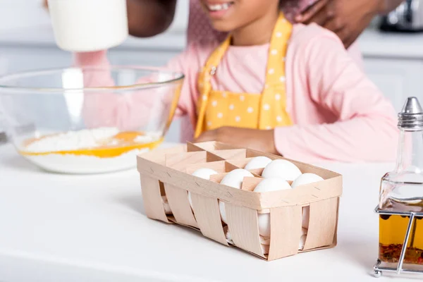Cropped view of african american kid in apron cooking on kitchen, eggs and oil standing on table on foreground — Stock Photo