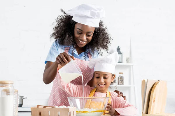 African american mother and daughter in chef hats pouring milk in bowl with ingredientes for dough — Stock Photo