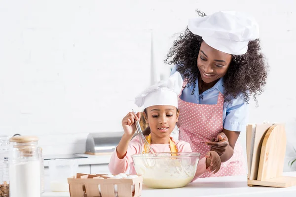 Mãe e filha africana americana em chapéus de chef misturando massa na cozinha com livro de receitas — Fotografia de Stock