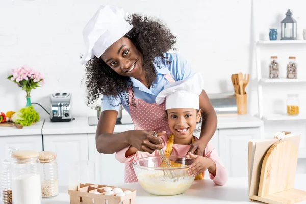 Afro-americana mãe e filha em chapéus chef misturando massa com batedor na cozinha — Fotografia de Stock